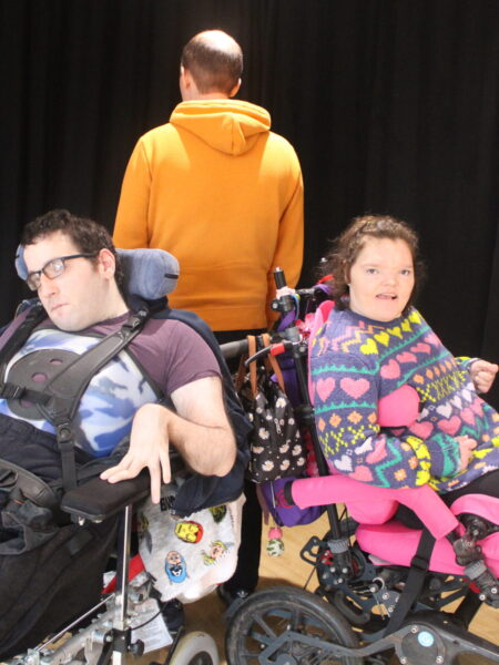 A man and woman each in wheelchairs and a man with his back to them. Enacting a performance.