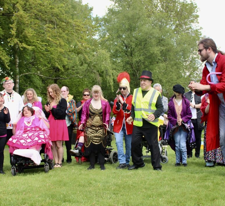 Group of disabled actors performing outside. A lady is in a gold dress , a man next to her wears a red cavalier jacket and helmet with red feathers. Another man is wearing a high vis jacket and a top hat.